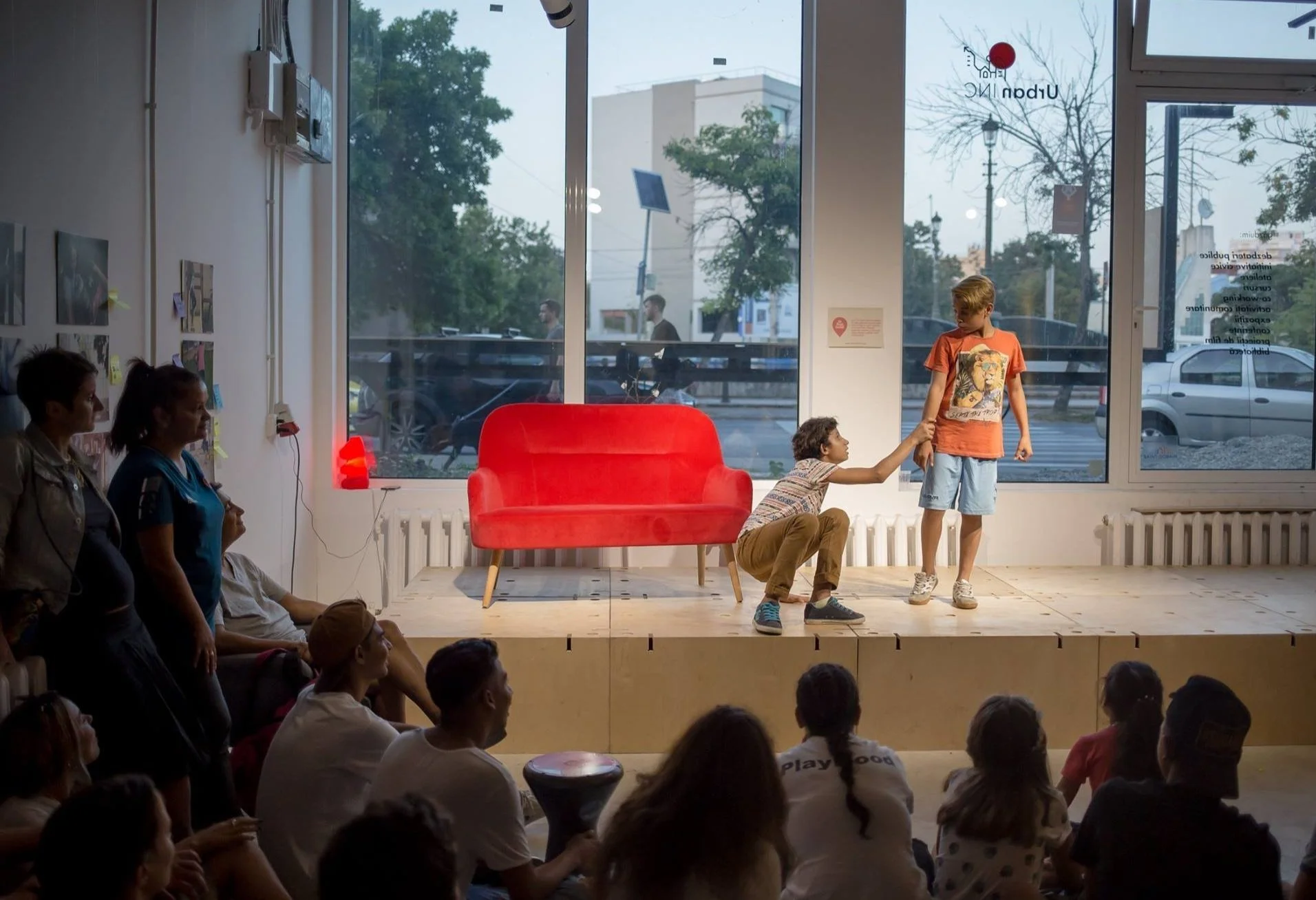 Poza din timpul unui spectacol de teatru susținut La Firul Ierbii. Doi copii se află pe scenă, unul dintre ei îl trage de mână pe celălalt. În jurul scenei se află mai multe persoane, atente la piesă