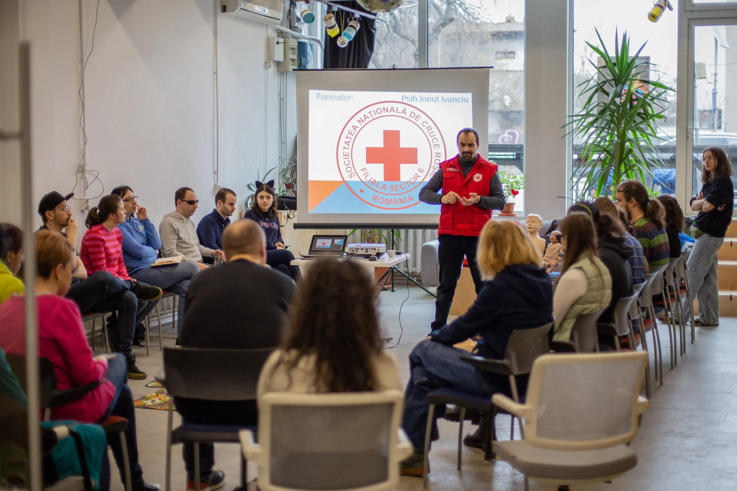 Poză surprinsă în timpul atelierului de prim ajutor. Pe un proiector vedem logo-ul Crucii Roșii. ALături, un bărbat îmbrăcat în roșu susține cursul în fața a multor persoane care stau în cerc