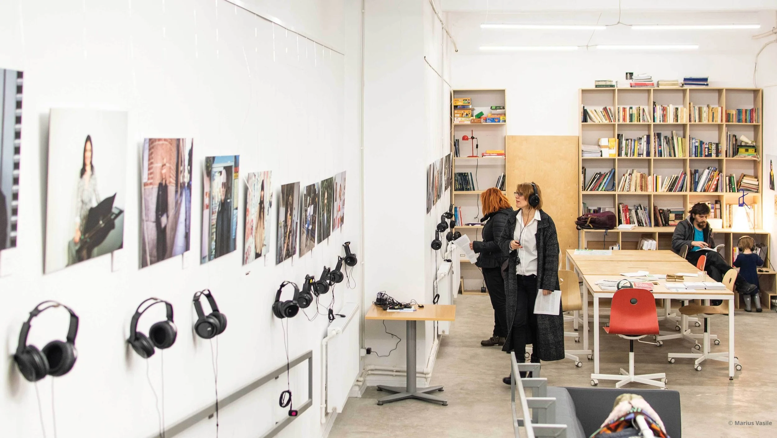 Imagine de la o expoziție din Biblioteca La Firul Ierbii. Pe pereți vedem poze și căști. Două persoane ascultă poveștile în timp ce privesc fotografiile
