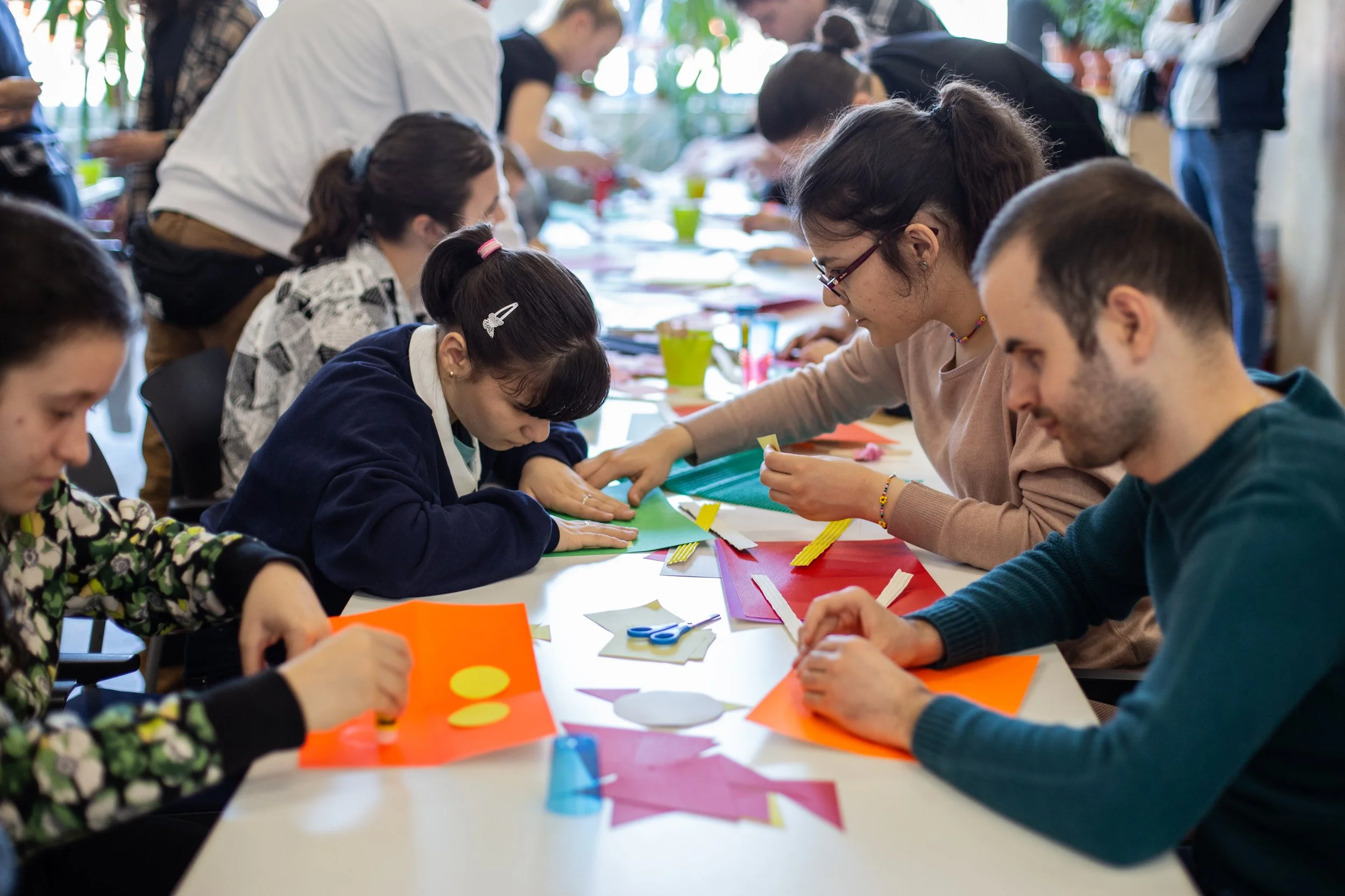 În imagine vedem o masă lungă. Mai multe persoane fac obiecte din origami